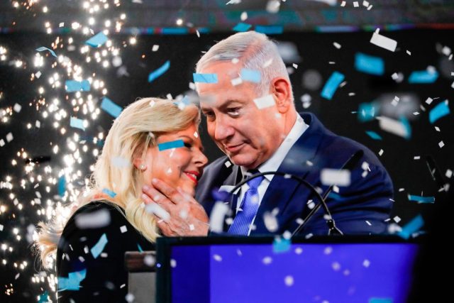 Netanyahu Likud Victory 2019 Corruption Benjamin Netanyahu speaks at a campaign event, surrounded by Likud party supporters holding blue and white flags.