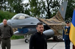 Zelensky Says Ukraine Receives First F-16 Jets This Year Ukrainian and Danish officials stand beside an F-16 fighter jet at a sunlit airbase press event.